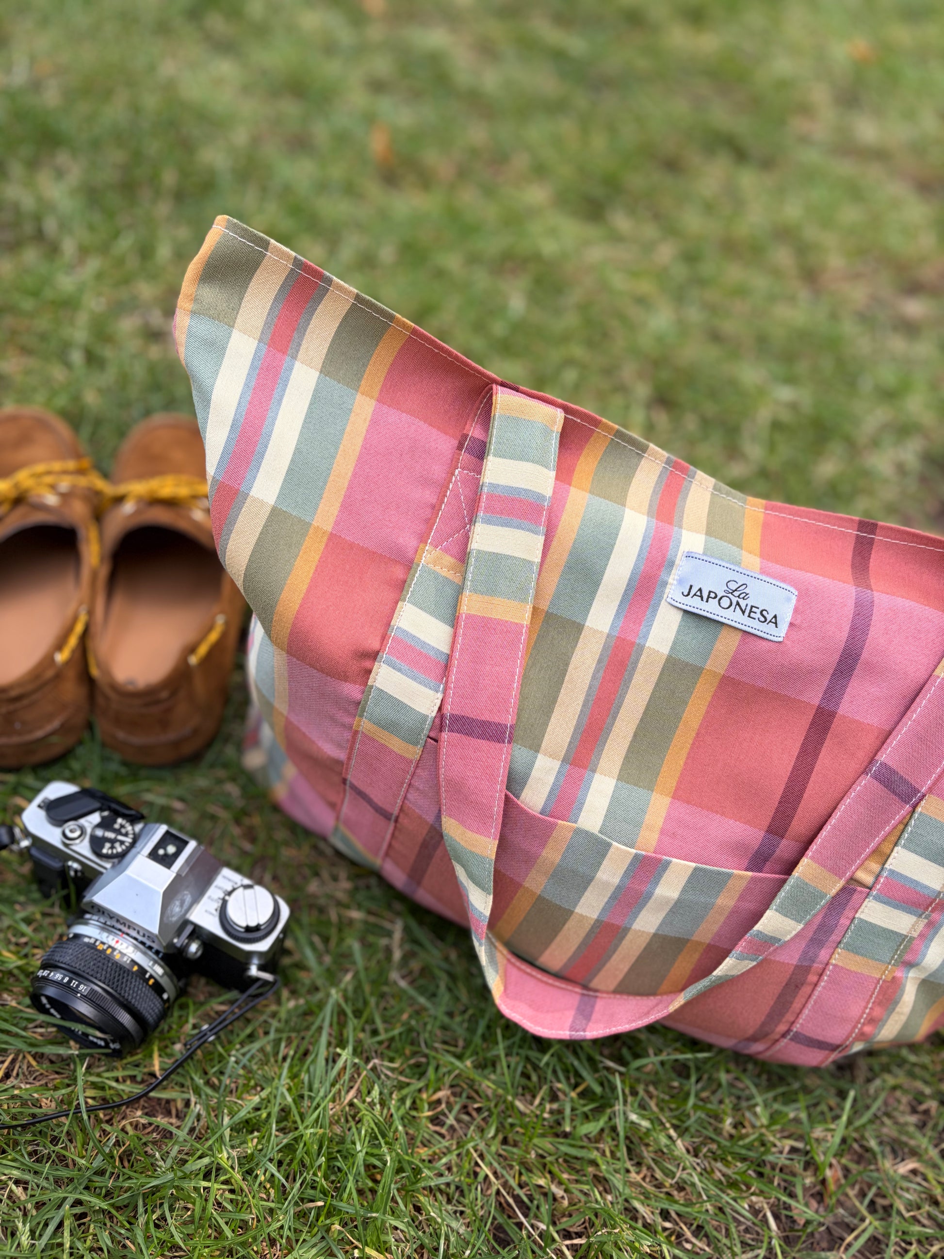 Tote bag tartán rosa sobre pasto junto a cámara analógica y zapatos