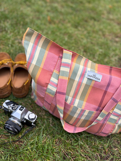 Tote bag tartán rosa sobre pasto junto a cámara analógica y zapatos